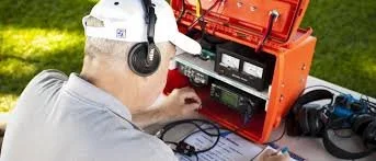 A person wearing headphones working with electronic equipment outdoors on a grassy area.
