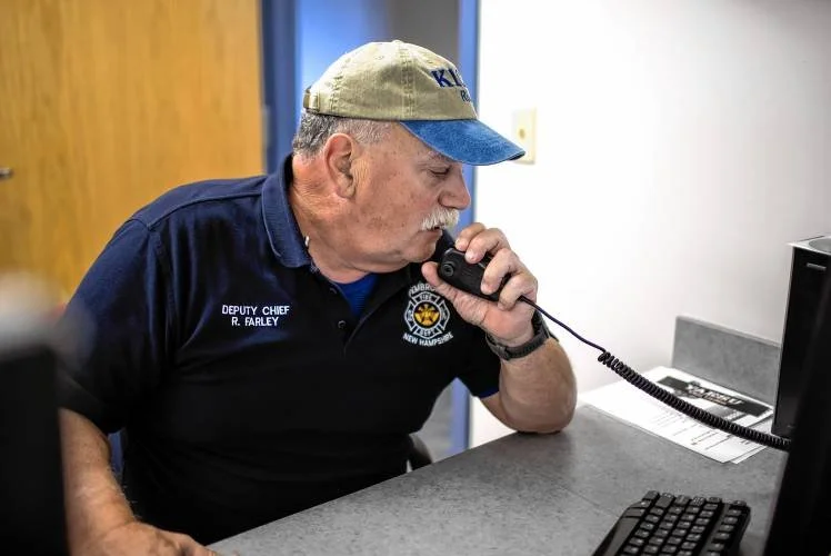A man wearing a navy uniform with a badge and a name tag, sitting at a desk and speaking on a landline phone. He has a gray mustache, a beige cap, and is looking at a computer monitor.