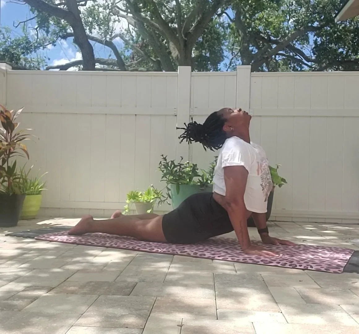 A woman practicing yoga outdoors on a pink and purple patterned mat, doing a cobra pose with her head tilted back, surrounded by potted plants and a white fence, with trees and a blue sky in the background.