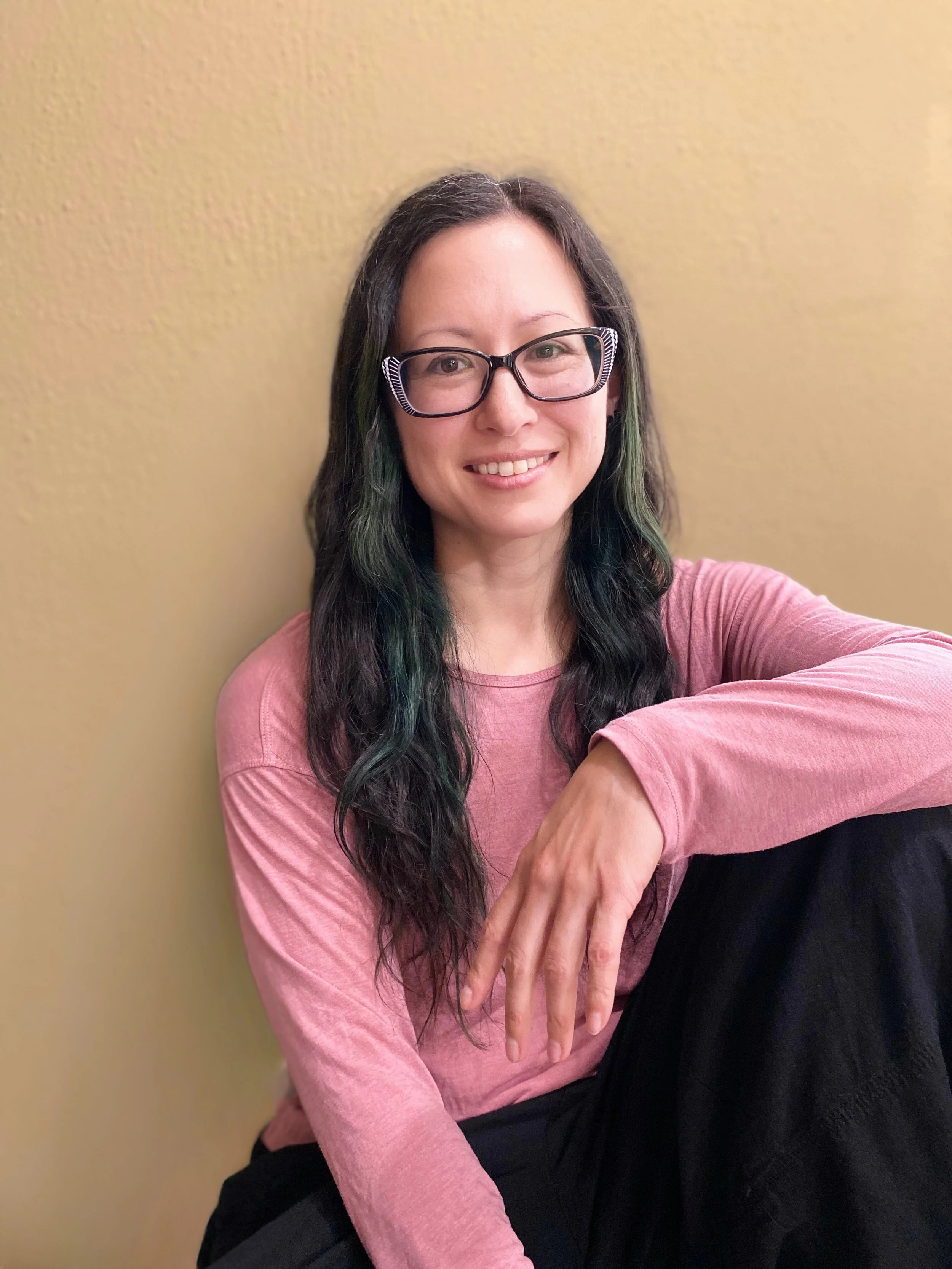 A woman with long dark hair with green highlights, wearing glasses and a pink long-sleeve shirt, sitting against a yellow wall and smiling at the camera.