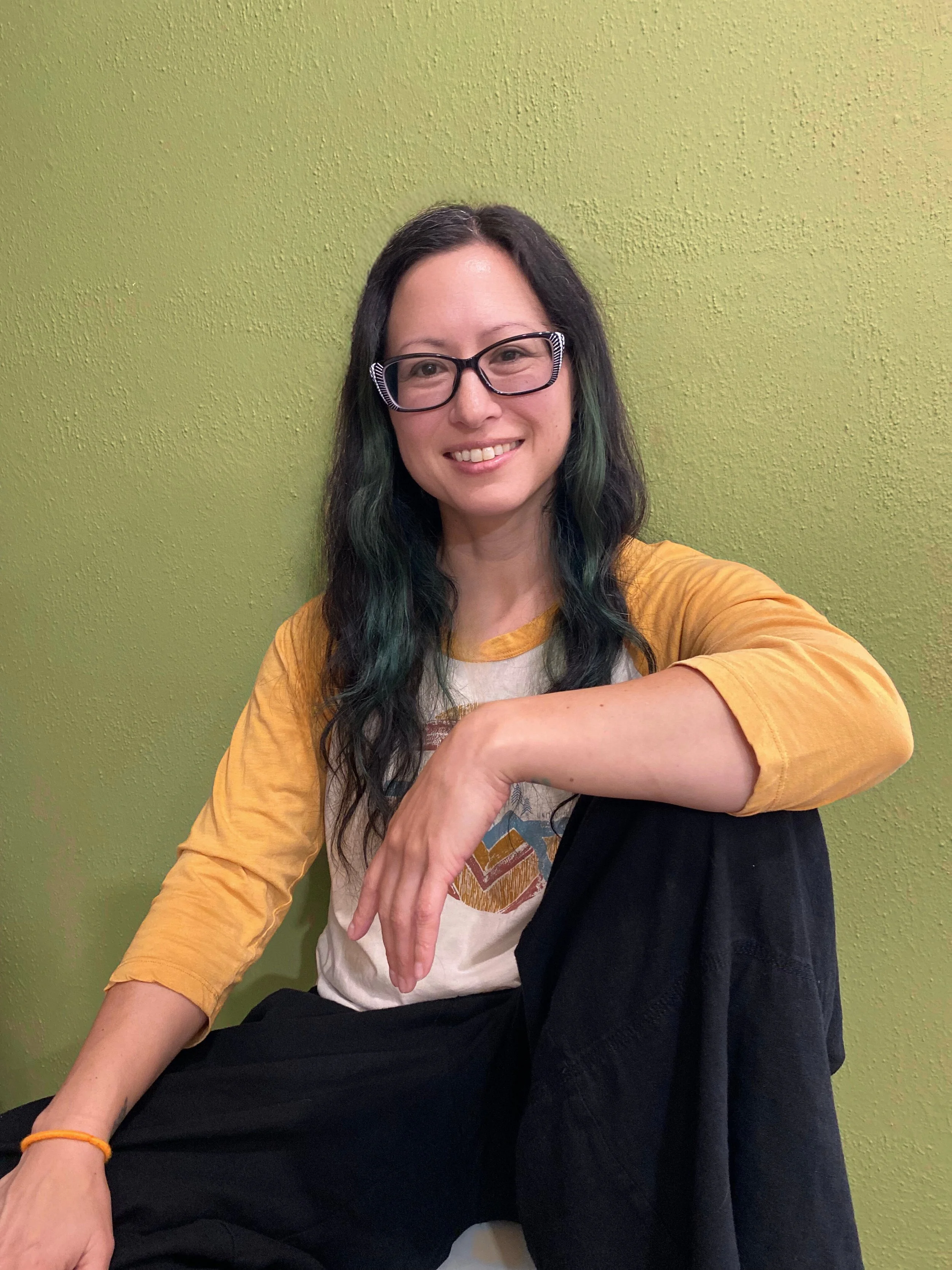 A woman wearing glasses and a yellow and white shirt, sitting against a green wall. She has long, dark hair with green highlights and is smiling at the camera.