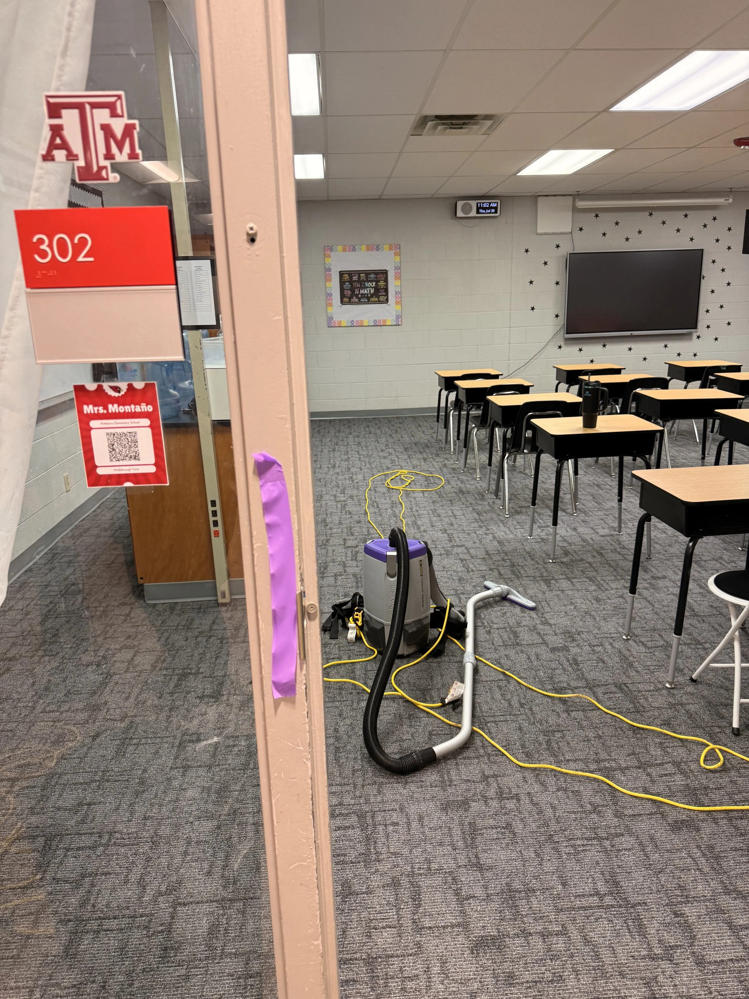 Classroom with desks and chairs, a large television, a mounted digital clock, and cleaning equipment on the floor, viewed through an open door.