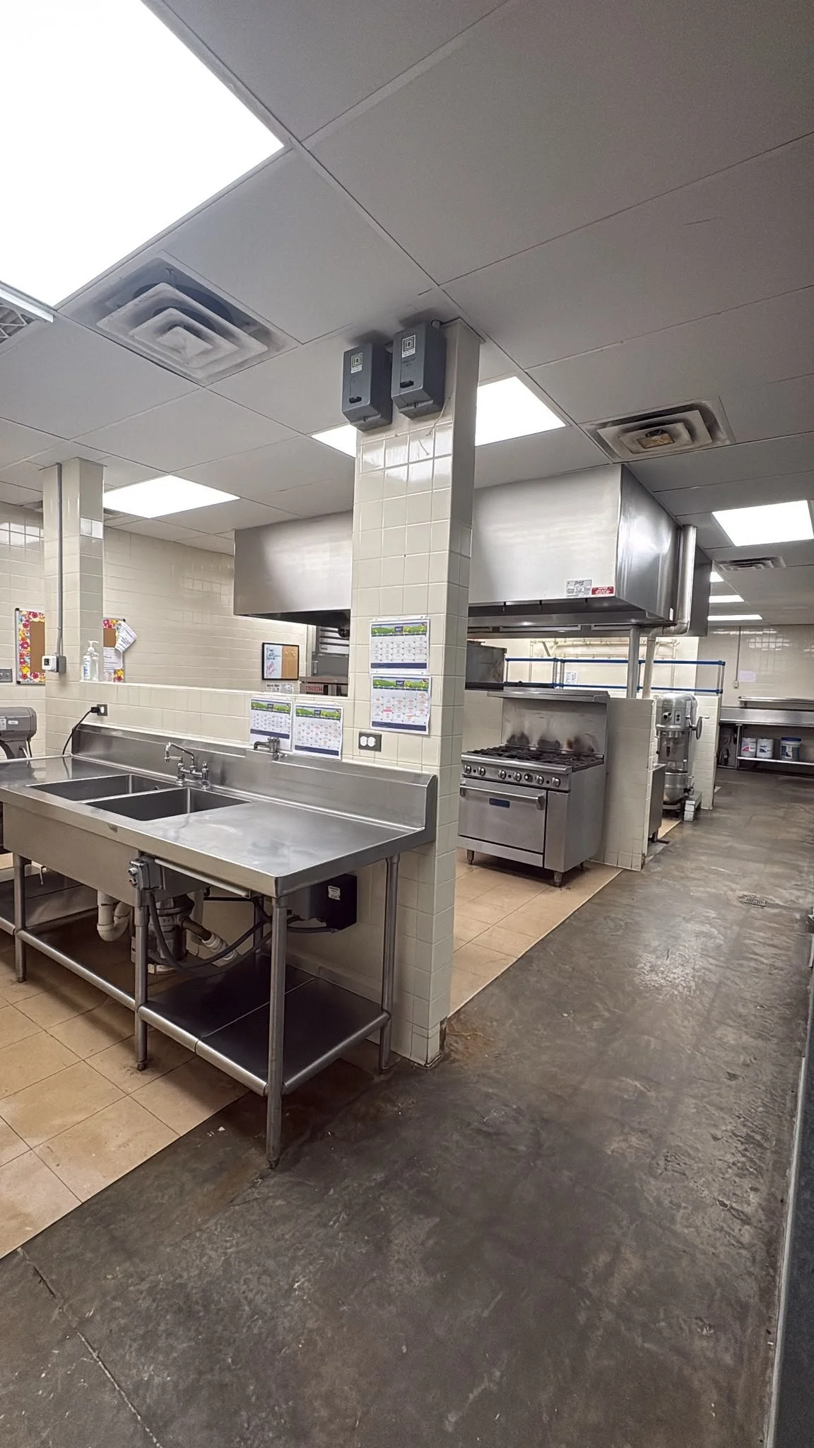 A commercial kitchen with stainless steel sinks, a stove, and wall-mounted calendars.