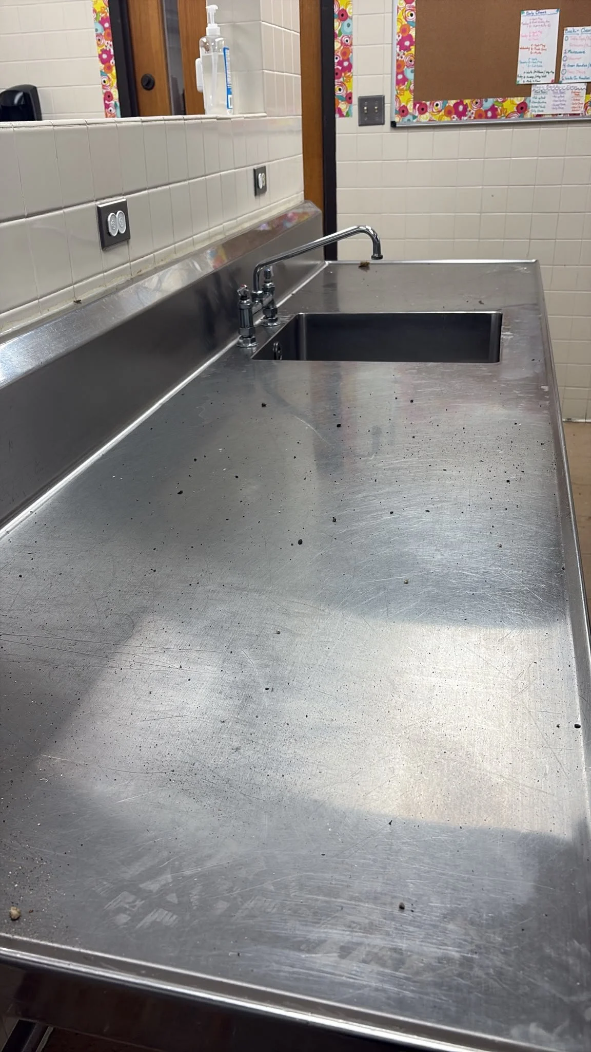 A stainless steel kitchen sink and countertop with some dirt and debris, in a room with tiled walls and a notice board.