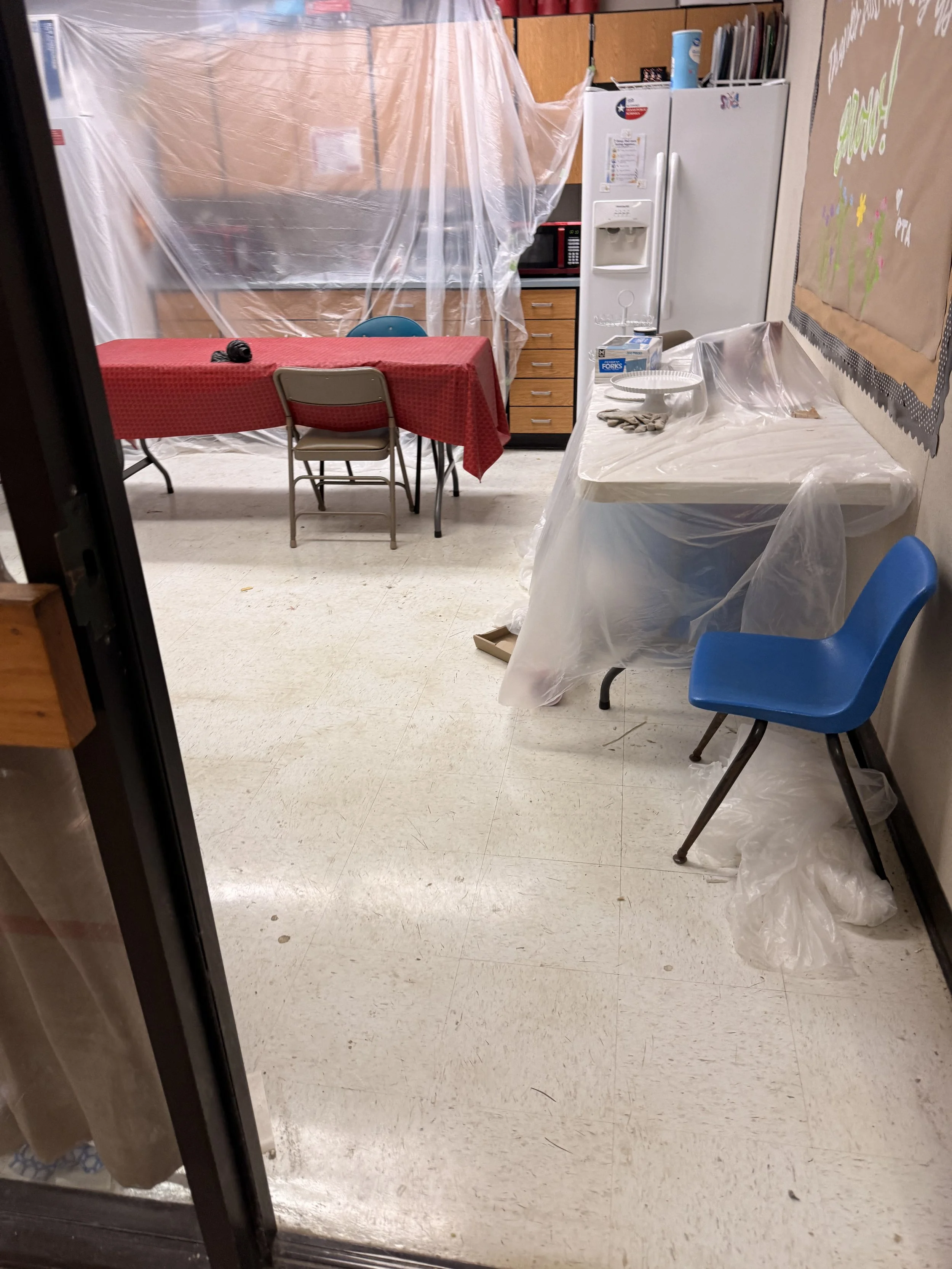 A room with a dining table covered with a red tablecloth, chairs, a refrigerator, a microwave, and a countertop with some items, all enclosed with plastic sheeting.