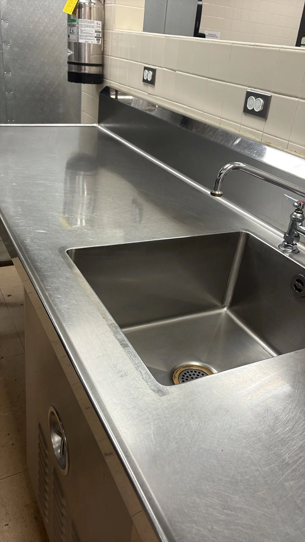 A stainless steel sink with a faucet and a drain, against a tiled wall with electrical outlets and a fire extinguisher in the background.