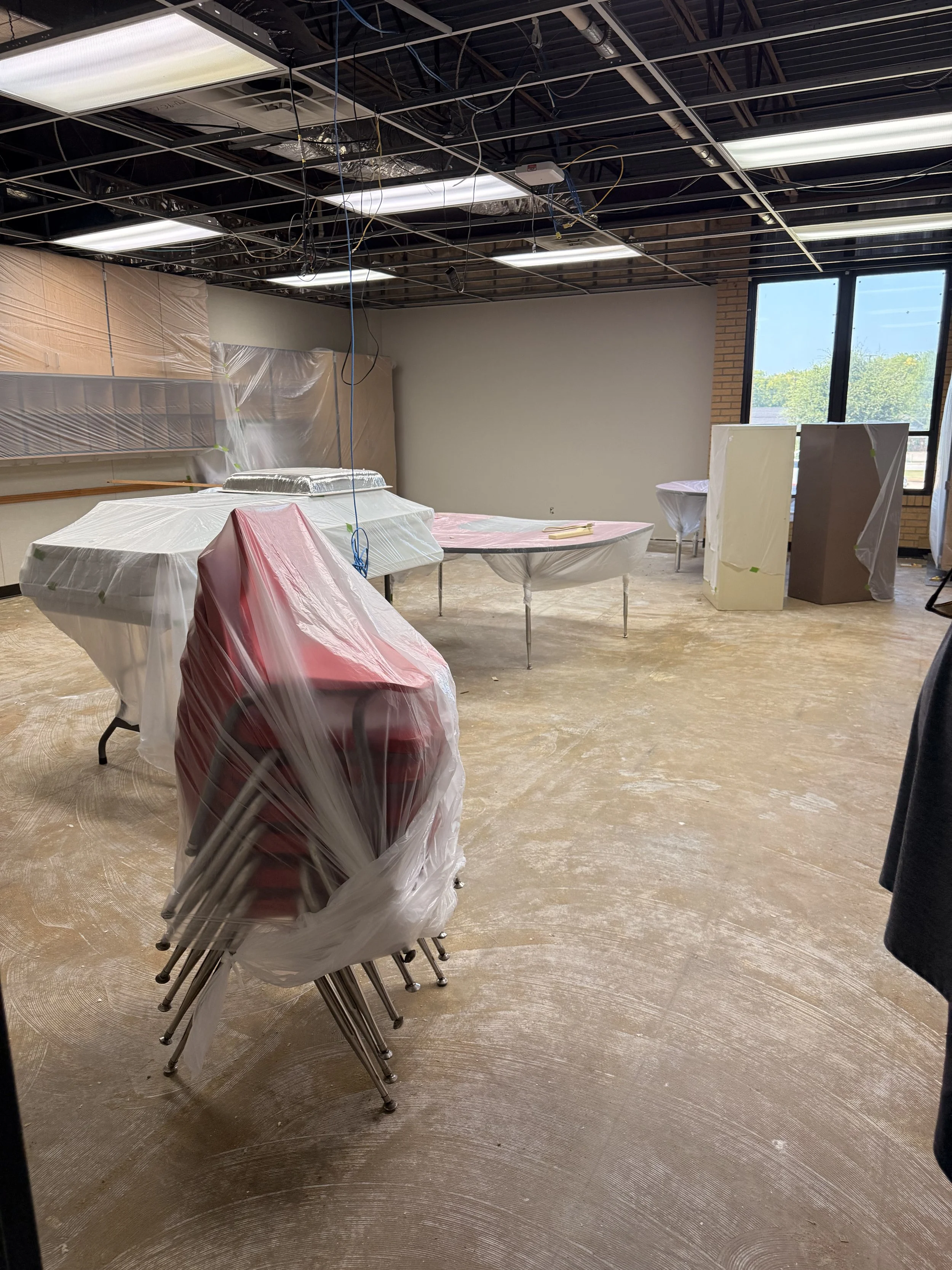 Empty room under renovation with tables covered in plastic, plastic sheeting, and exposed ceiling with wiring, large window showing trees outside.