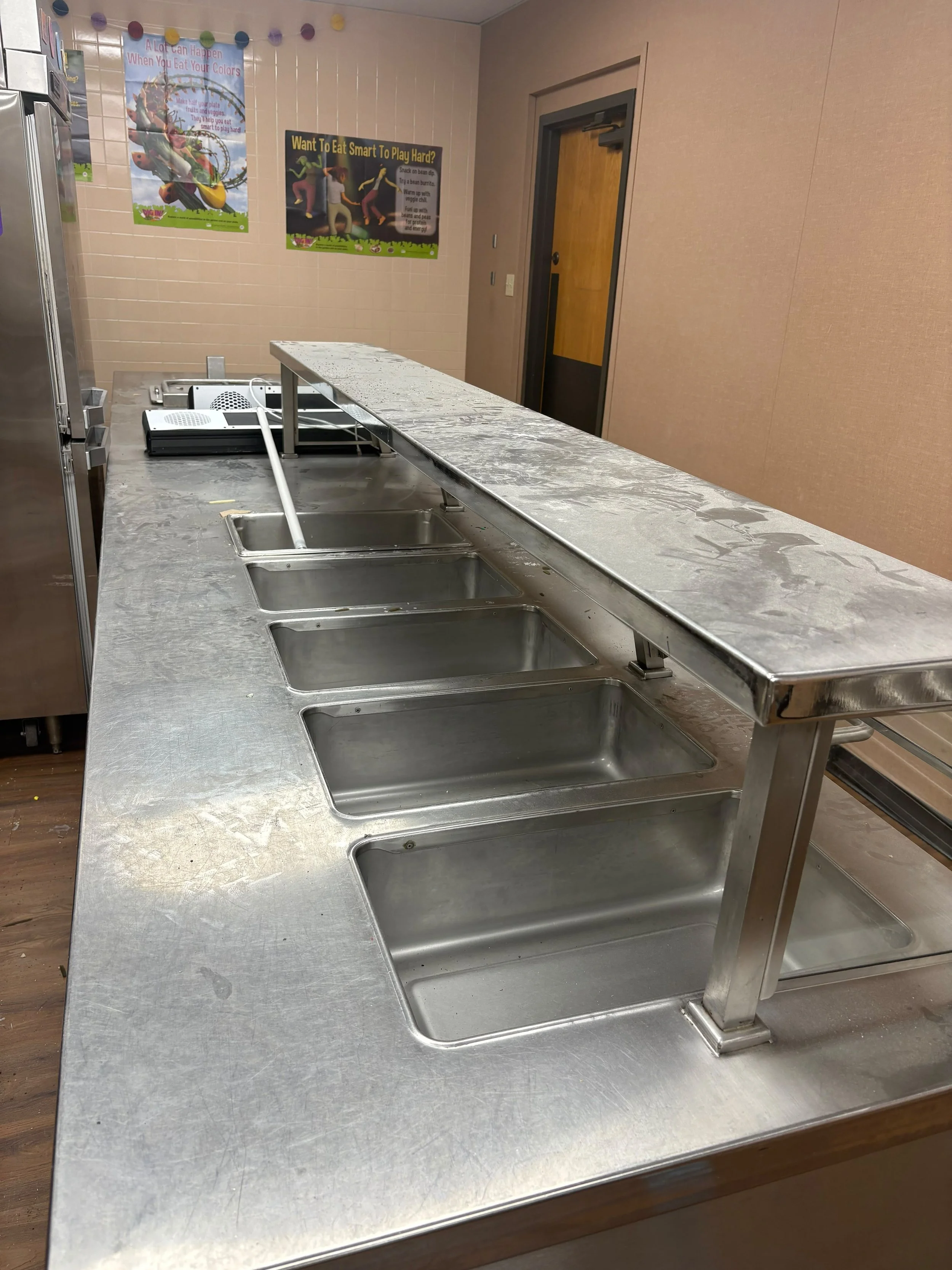 A stainless steel countertop with four empty sinks in a room with pink walls. Two posters are on the wall, one with the text 'A Lot Can Happen When You Eat Your Colors' and the other with the text 'Want To Eat Smart To Play Hard?'