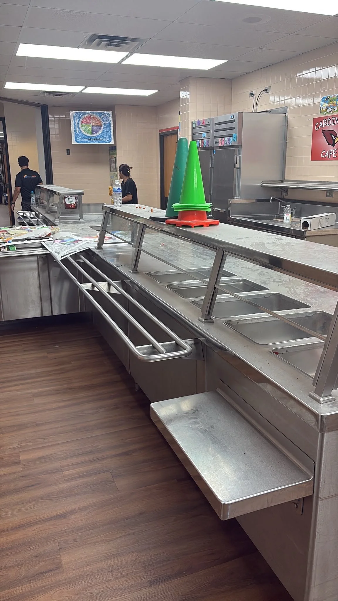 Empty cafeteria salad bar with green plastic cones on the counter, and two staff members in background.