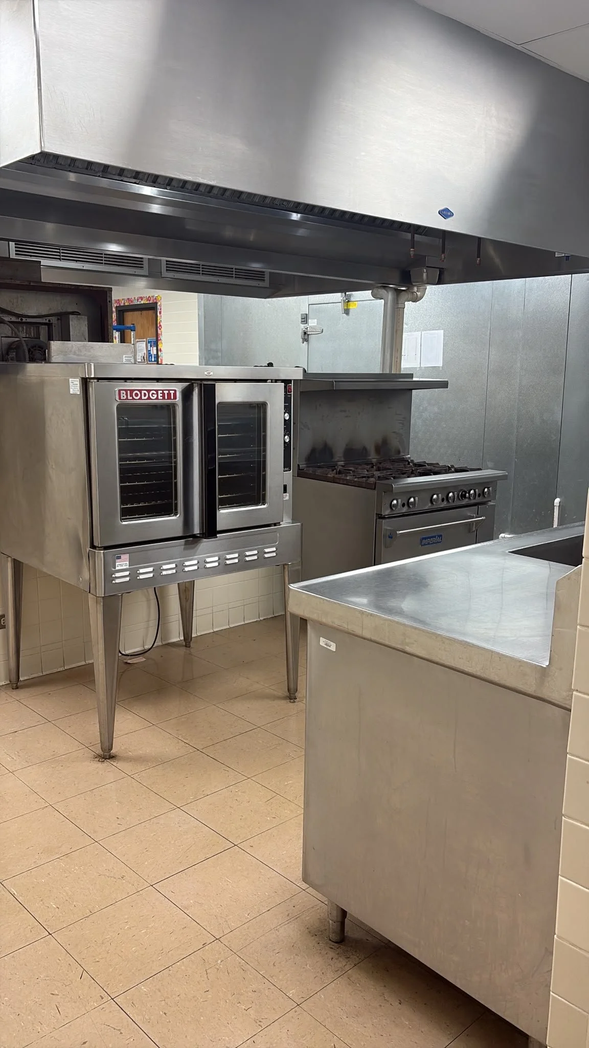 Commercial kitchen with stainless steel appliances, including an oven, stove, and a metal table with a rounded corner, beige tiled floor, gray walls, and a large stainless steel vent hood overhead.