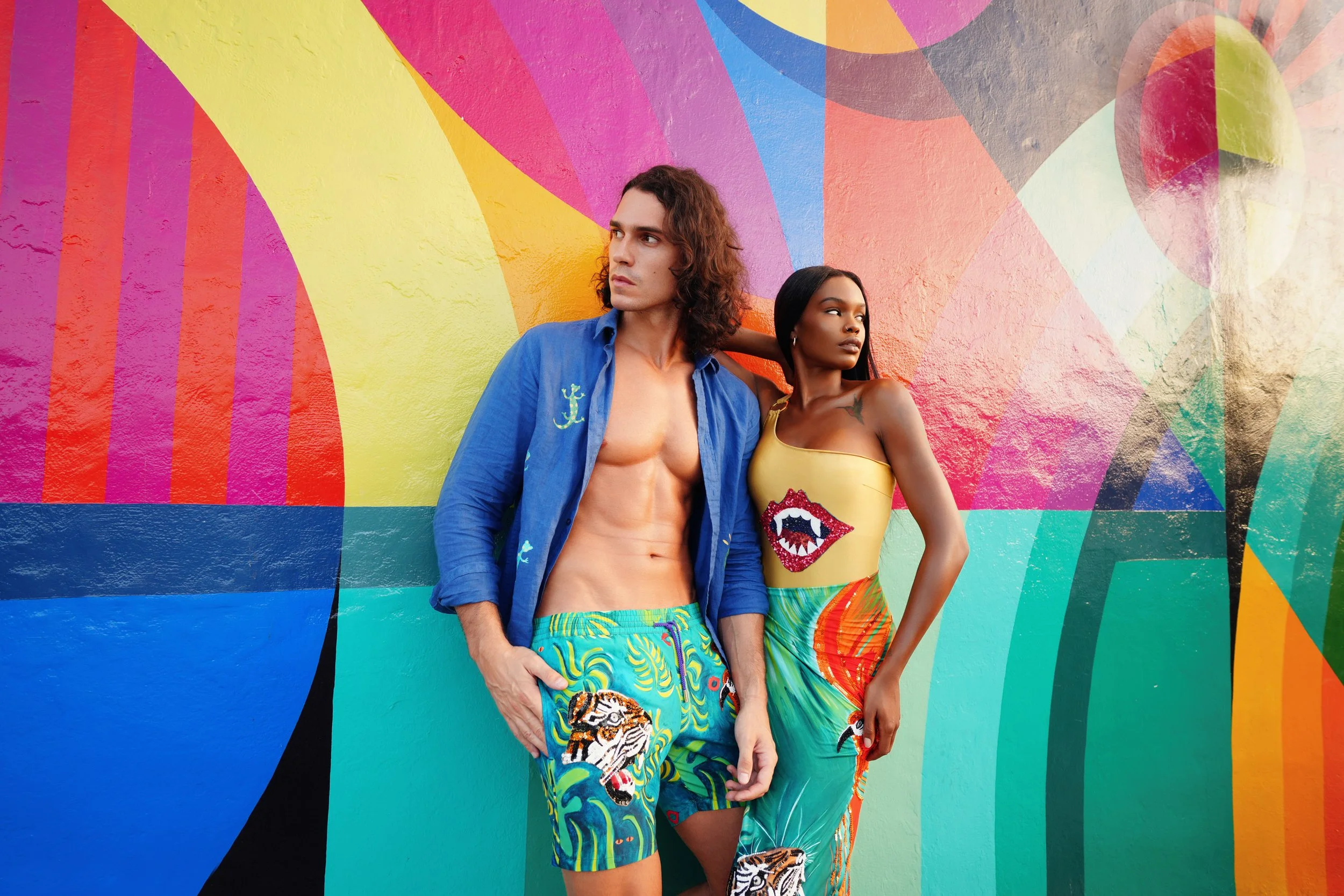 man and woman standing in front of a painted colorful wall in Miami Wynwood art district. The man wears a hand painted linen blue shirt with blue, green and yellow jungle swim shorts. The woman wears a one-piece asymmetrical gold swimming-suit
