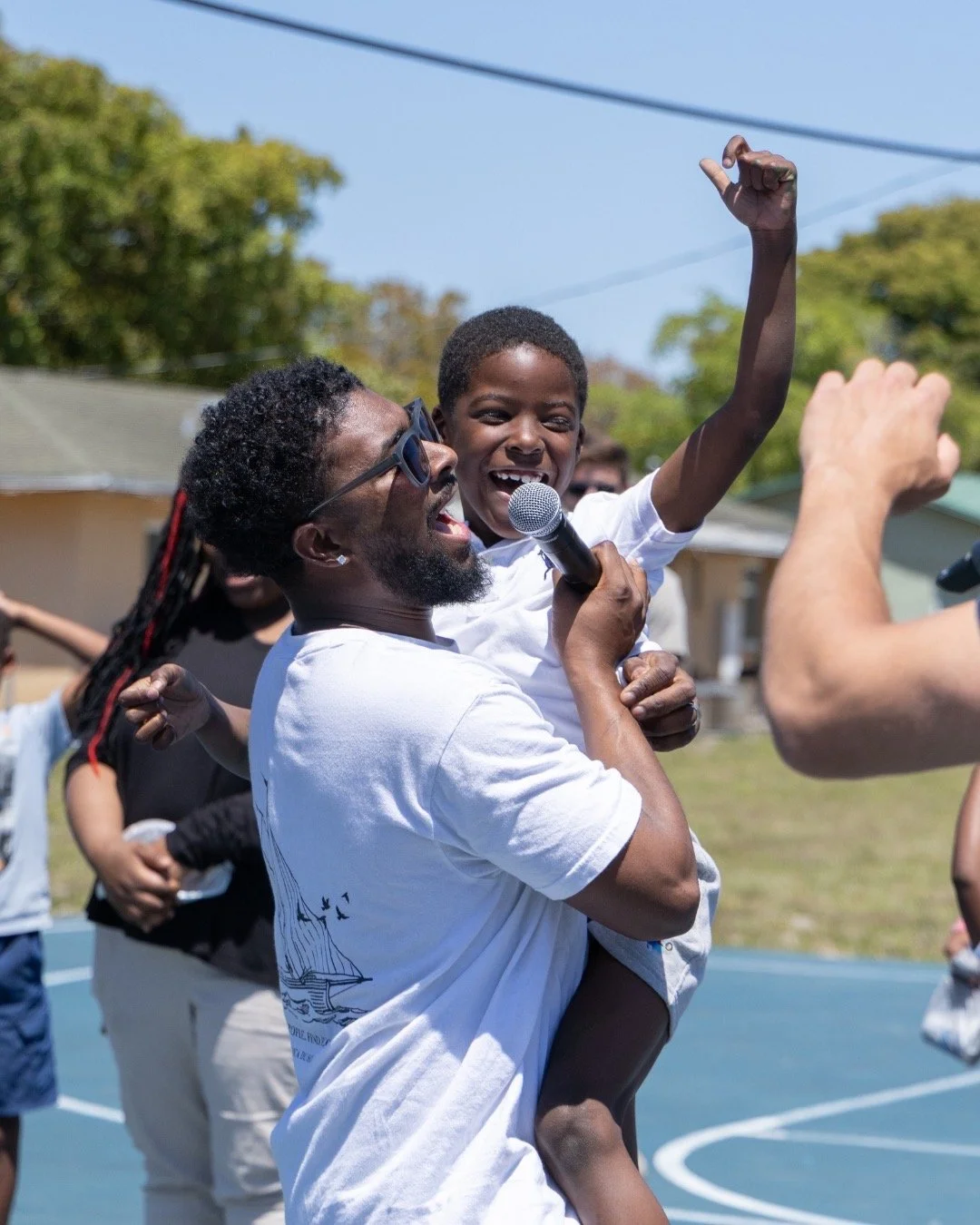 Man and boy celebrating together outdoors on a sunny day, with the man holding the boy in his arms and the boy smiling and raising his fist, while holding a microphone.