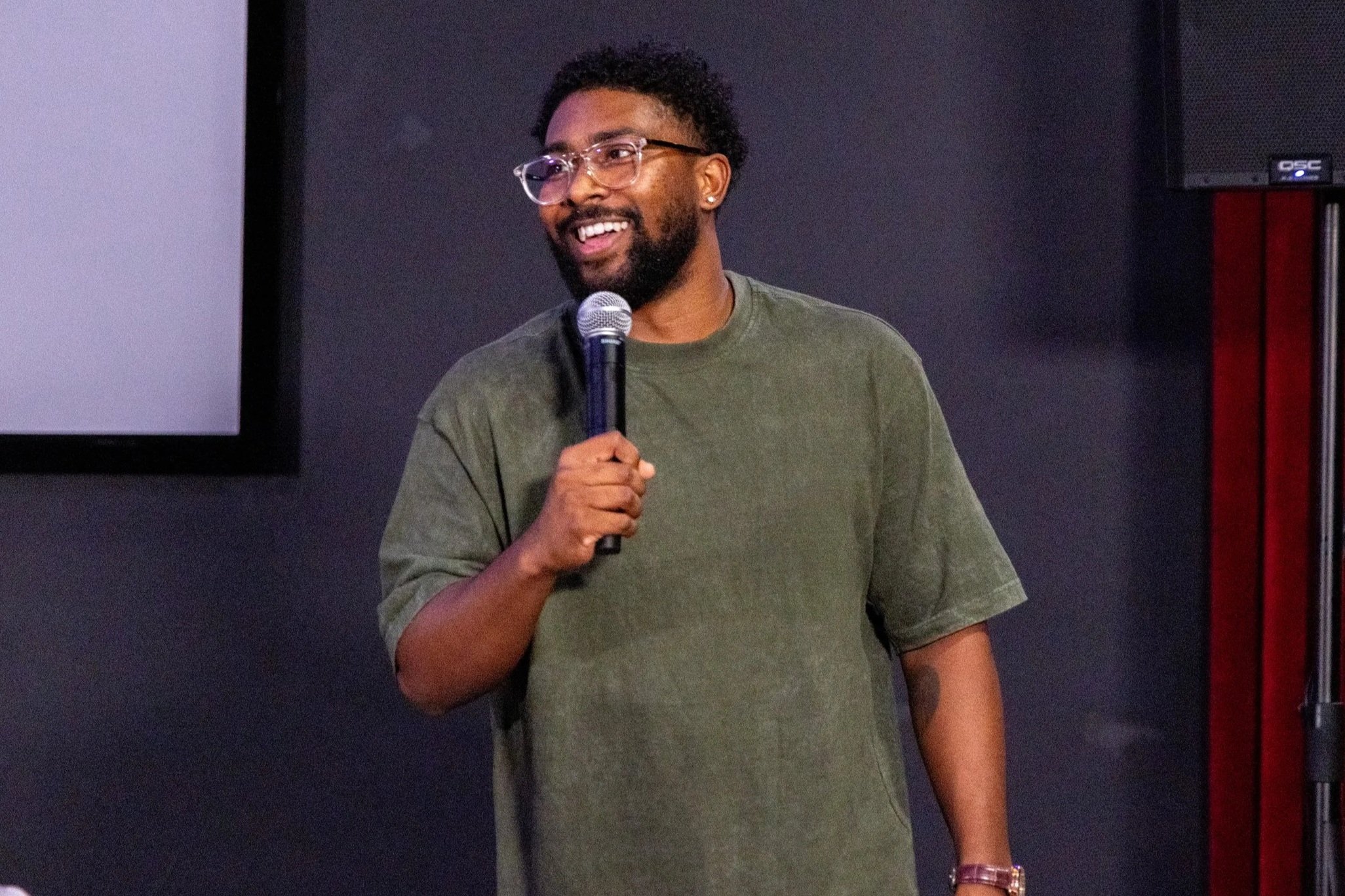 A man with glasses, a beard, and curly hair holding a microphone, smiling, standing indoors against a dark wall.