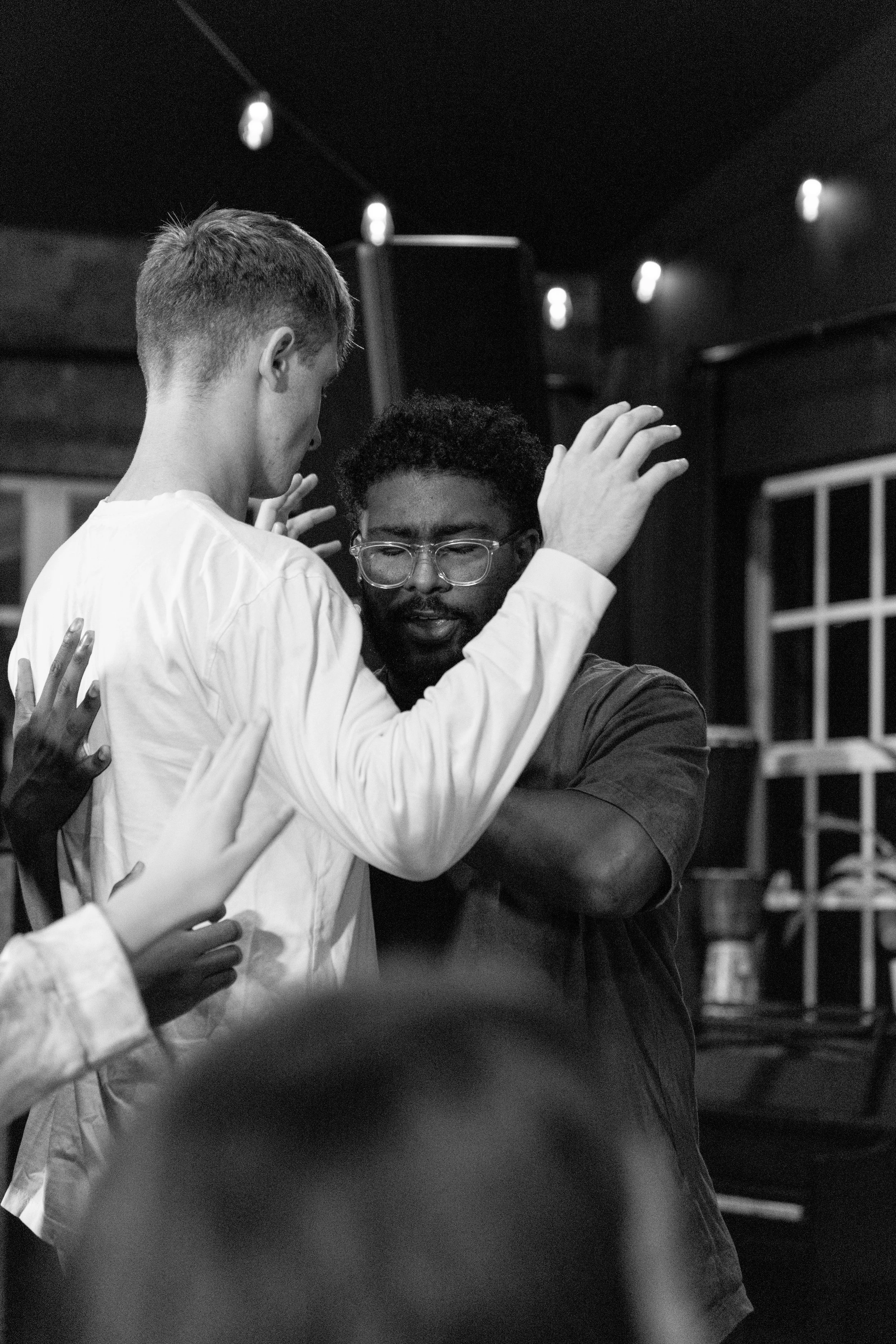Two men dancing closely, one with glasses and curly hair, wearing a dark shirt, and the other with short hair, wearing a light shirt, in a dimly lit room.
