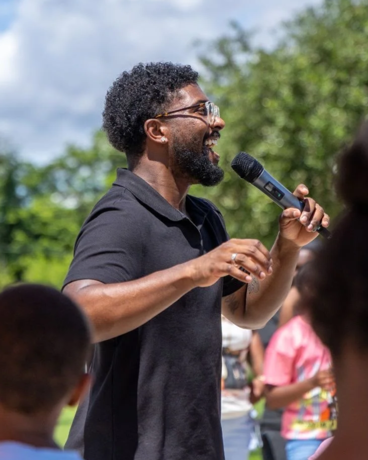 A man with glasses and a beard speaking into a microphone outdoors during daytime, with children and trees in the background.