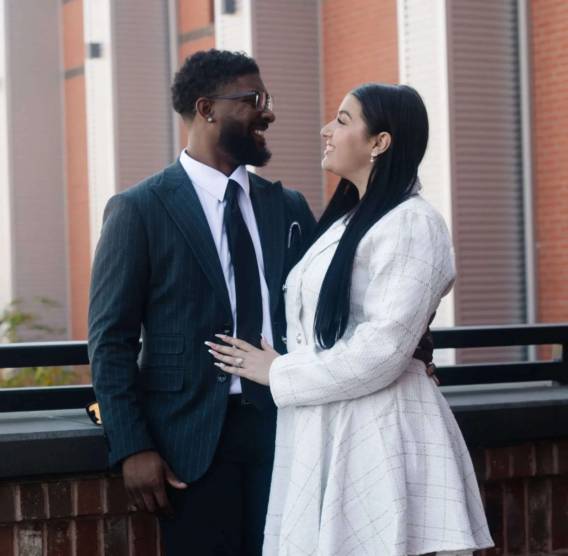 A couple dressed in formal attire smiling at each other outdoors, with the man in a dark pinstripe suit and the woman in a light-colored coat.