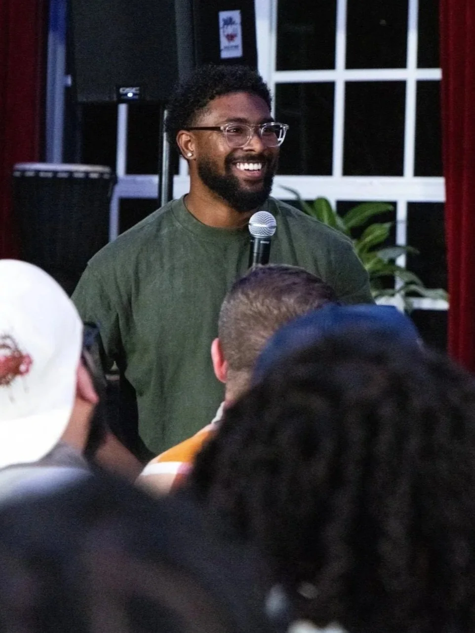 A man with glasses and a beard, smiling, holding a microphone, speaking to an audience in a room with window and plants in the background.