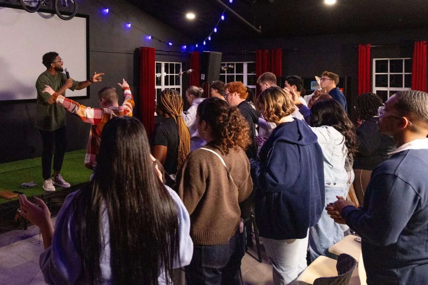 A speaker holds a microphone and points at an audience during a presentation or event in a dark room with string lights and red curtains.