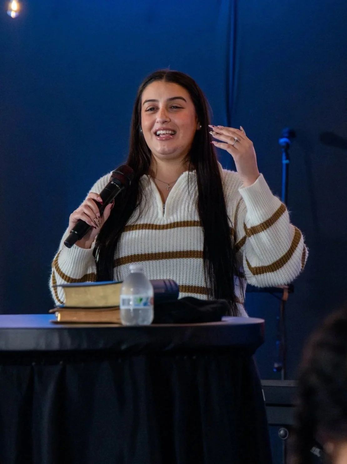 A woman with long dark hair speaking into a microphone at a table with books, a water bottle, and papers, in a dimly lit room with a blue wall background.