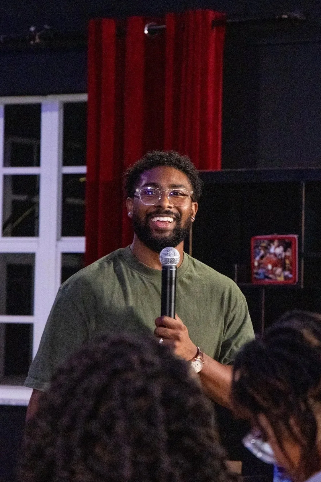 A man with glasses, a beard, and curly hair holding a microphone and speaking in front of an audience.