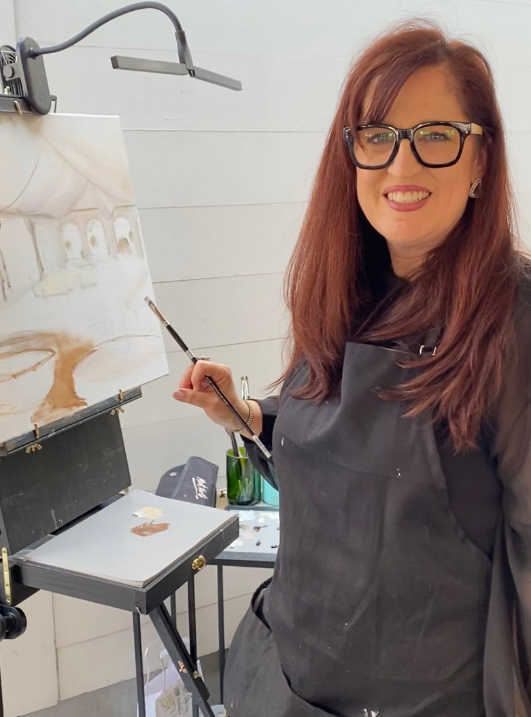 A woman with long red hair, glasses, and a black apron holding a paintbrush in front of an easel, painting a landscape scene in a bright, white room.