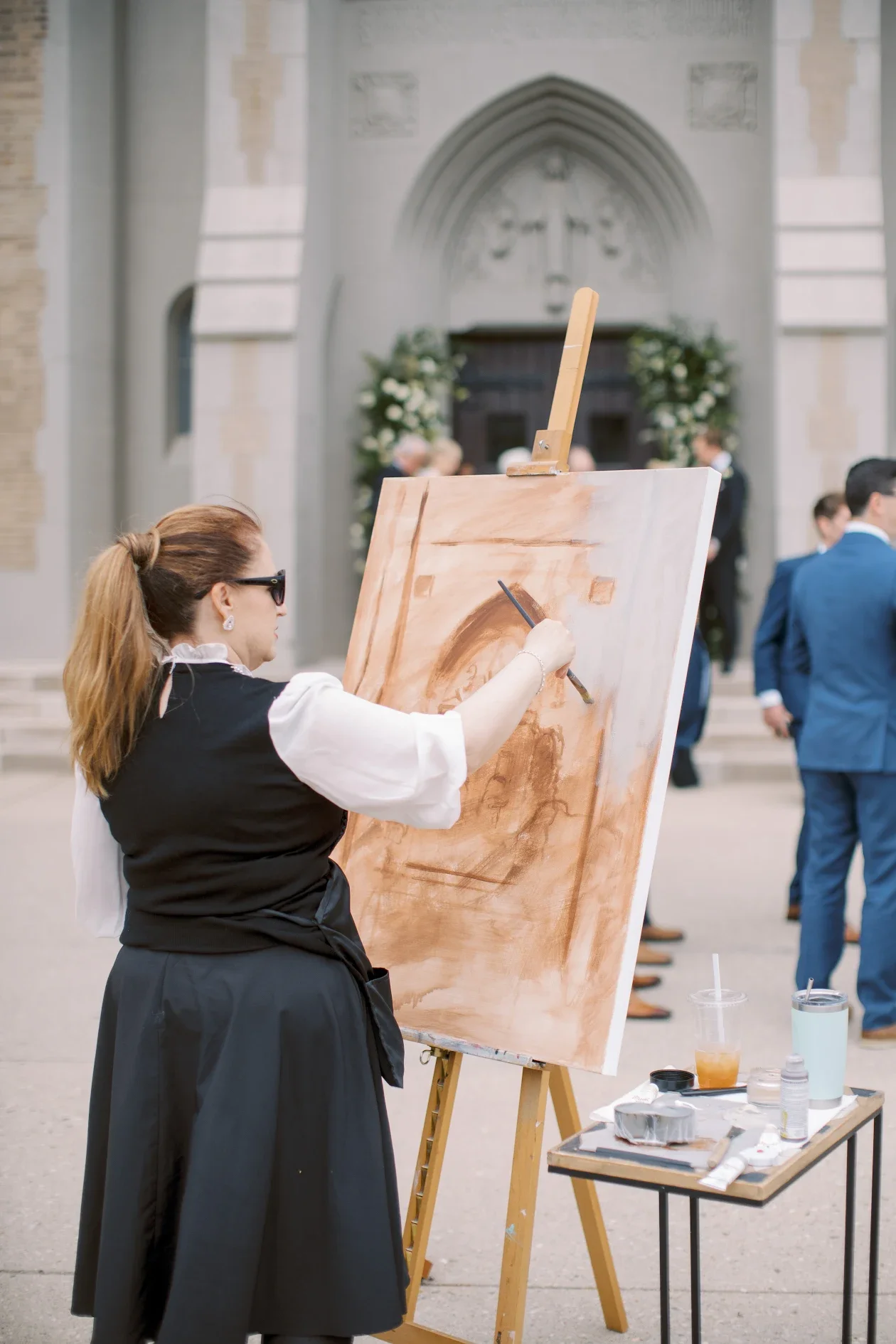 A woman in sunglasses and a black and white dress paints on a large canvas set up on an easel outside in front of a church. The woman is working on a detailed painting of an architectural structure with an archway. There are people in formal attire gathered around in the background.