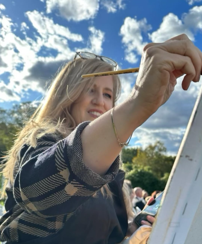 A young woman with long blonde hair is painting outdoors on a white canvas or easel, holding a yellow paintbrush. She is wearing a plaid shirt and glasses on her head, with a bright, partly cloudy sky behind her.