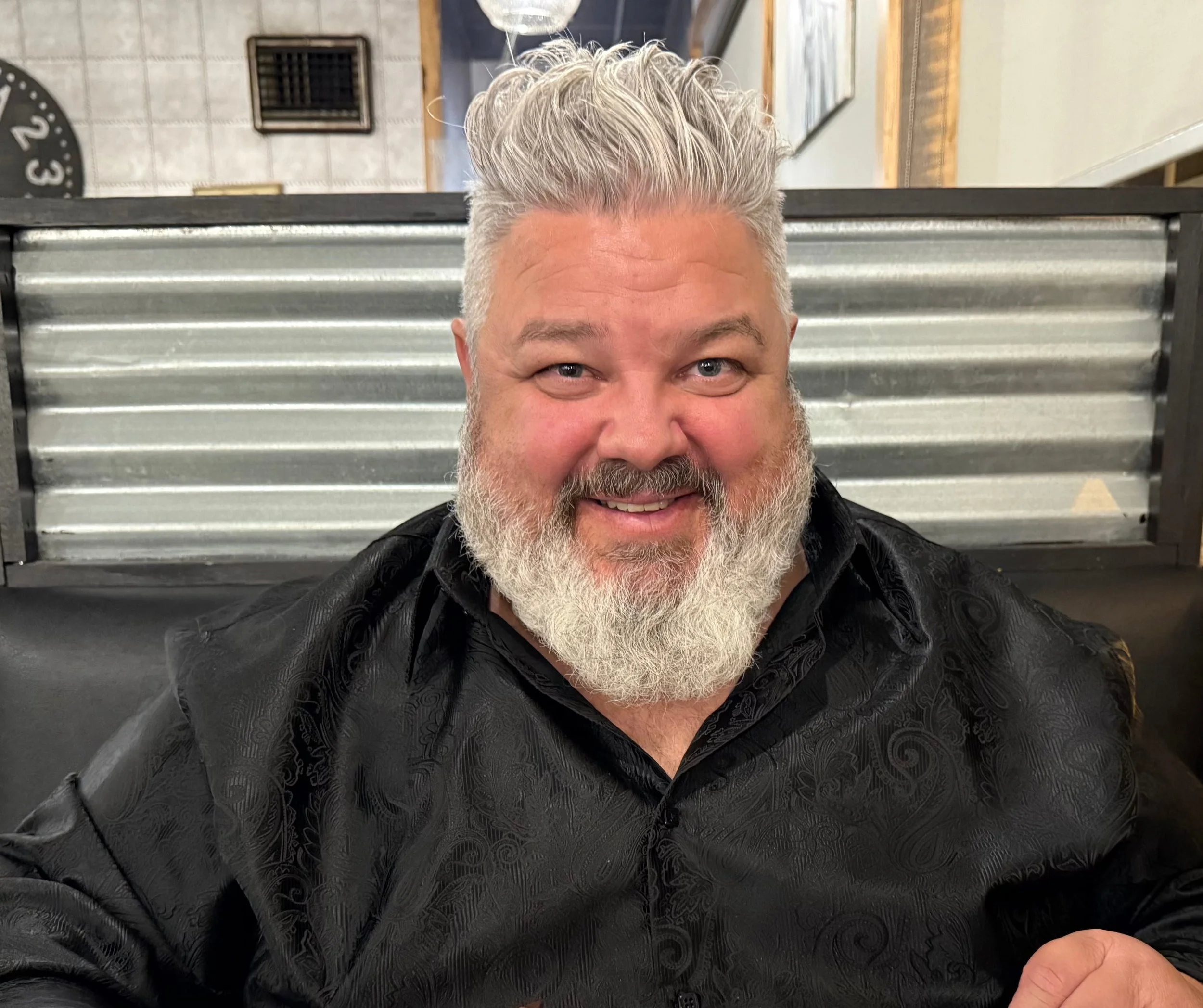 A smiling man with a gray beard and gray hair, wearing a black patterned shirt, seated indoors with a metal decorative wall panel and a room with framed pictures in the background.
