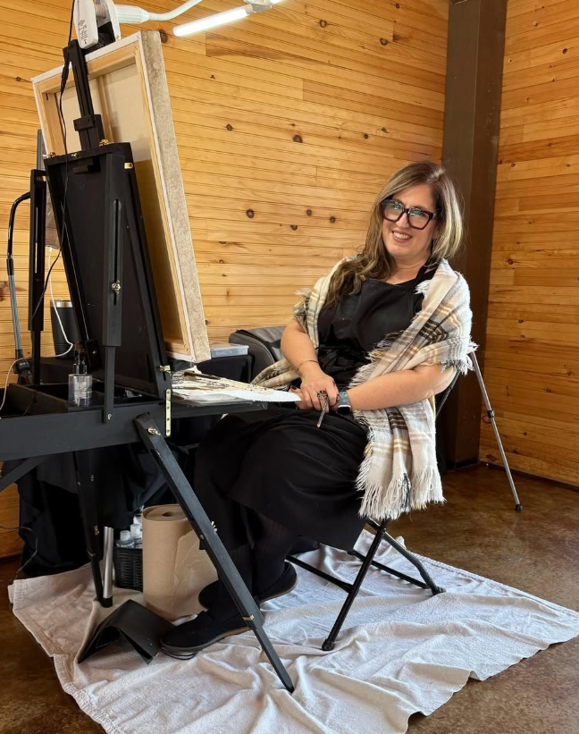 A woman with glasses sitting at an easel in a wooden art studio, smiling and holding a painting tool, wearing a black apron and a plaid shawl, with art supplies and paper towels on her table.