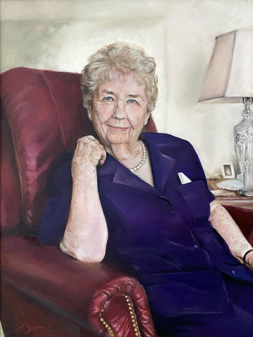 A senior woman with curly gray hair sitting in a red armchair, wearing a purple blouse and pearl necklace, showing a gentle smile in a living room with a table lamp and framed photo in the background.