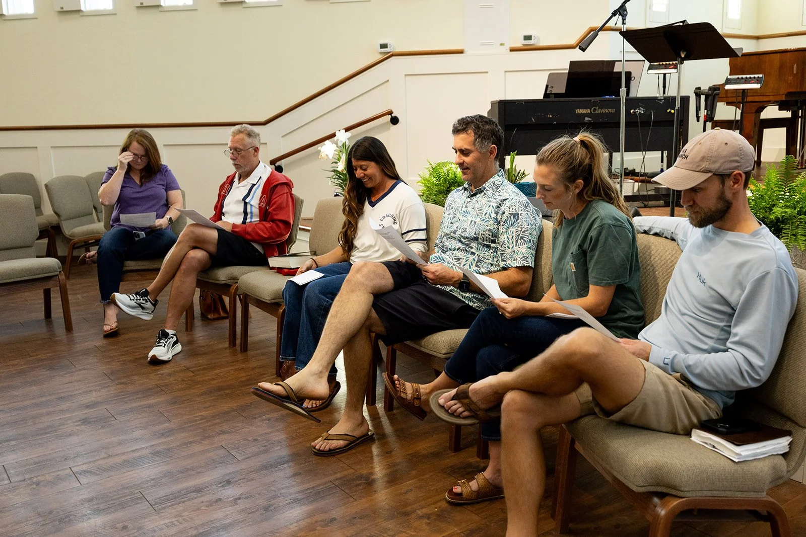 Six people are sitting on chairs in a room, looking at papers or books, with a music stand and musical instruments in the background.