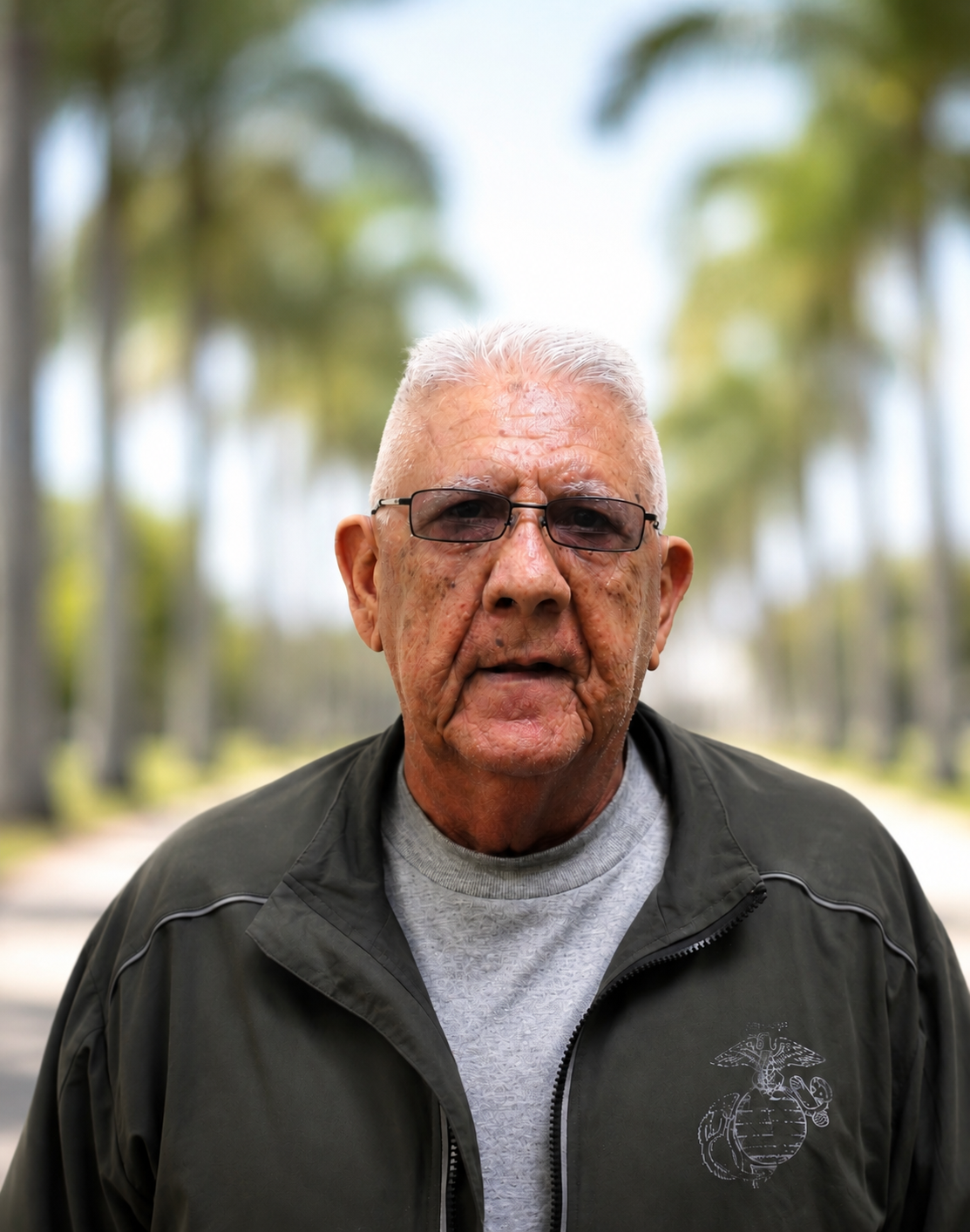 PBIC Staff member, an elderly man with gray hair, glasses, and textured skin, wearing a gray t-shirt and a dark gray jacket with a military emblem, standing outdoors with blurred trees and a light sky in the background.