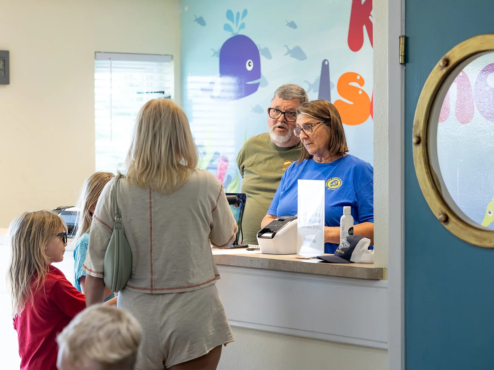 People at a service counter, including a woman in a light-colored shirt speaking to staff, surrounded by children, with colorful sea creature wall art in the background.