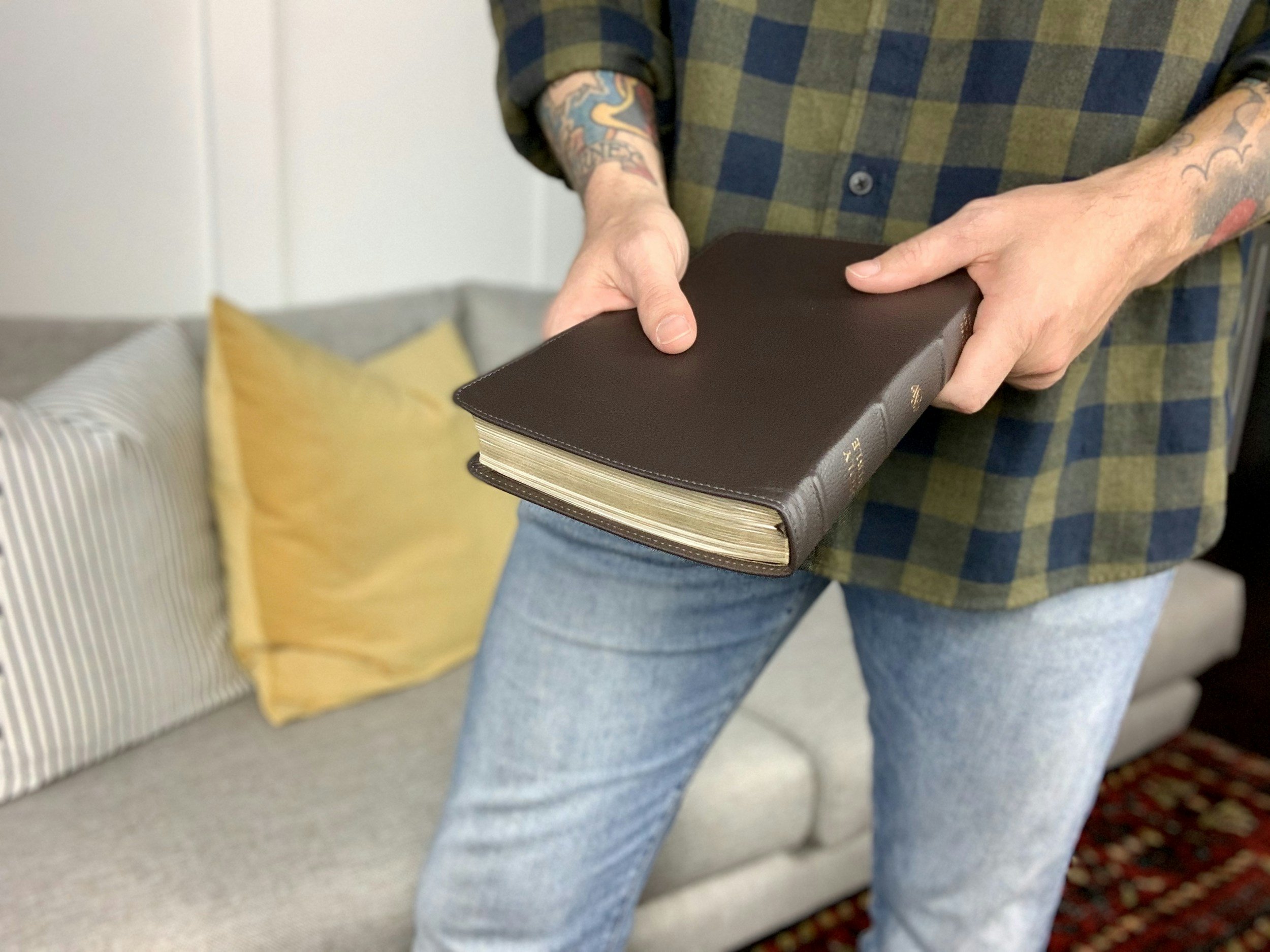 A person with tattoos holding a closed leather-bound book or journal in a living room.