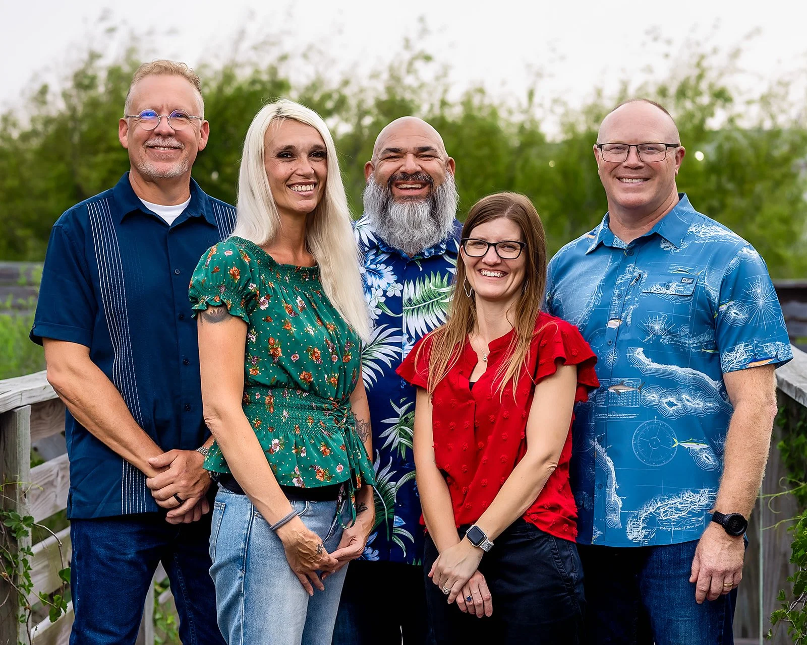 The staff of Padre Island Baptist Church: Group of five smiling adults standing outdoors on a wooden walkway with green trees in the background.
