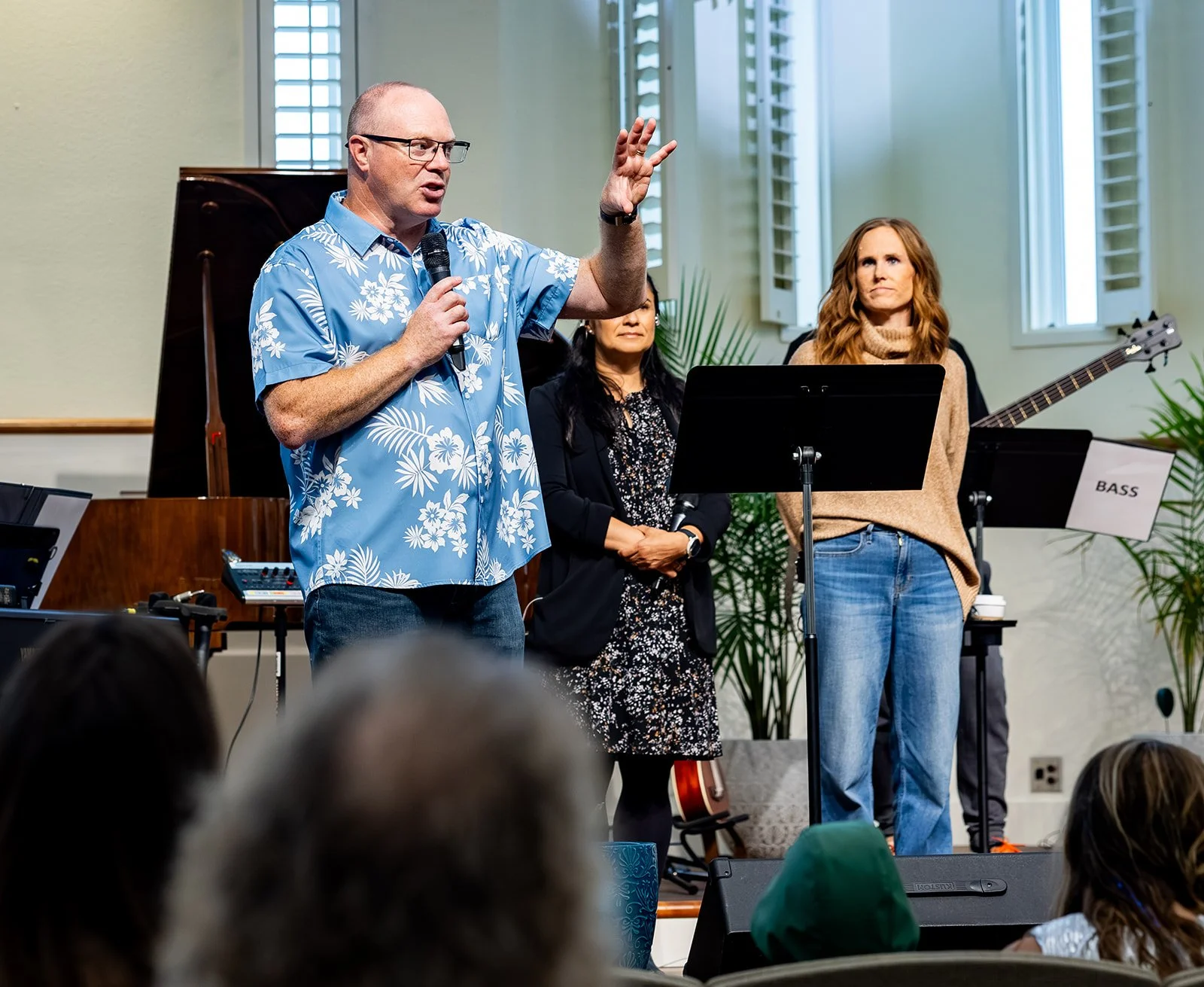 Family Pastor Steve of PIBC, in a blue Hawaiian shirt speaking into a microphone during a performance or presentation, with two women standing behind him; one with long dark hair and the other with long red hair holding a guitar.