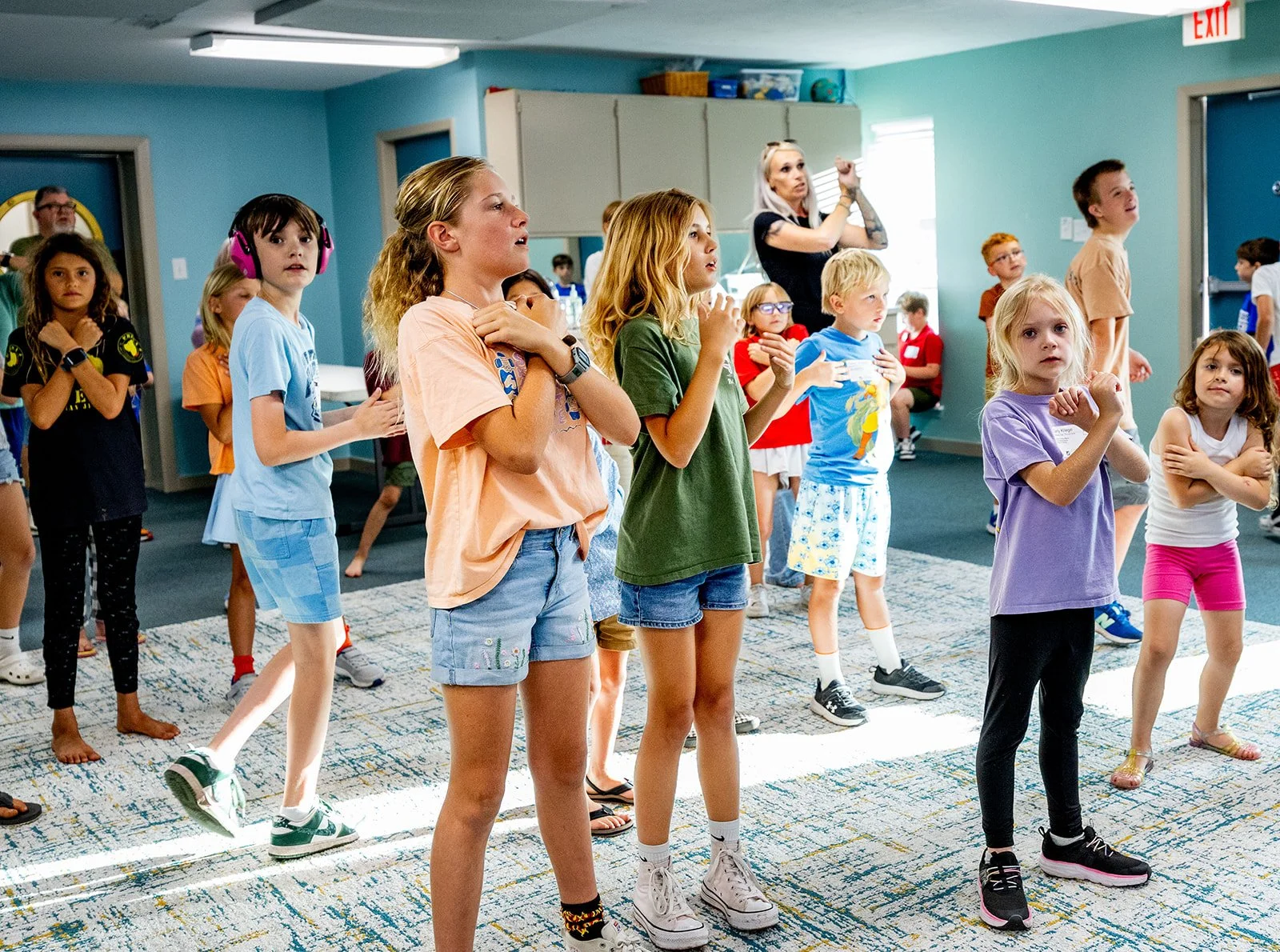 Group of children and a woman inside a room, some children are standing and appear to be engaged or singing, in a classroom or daycare setting.