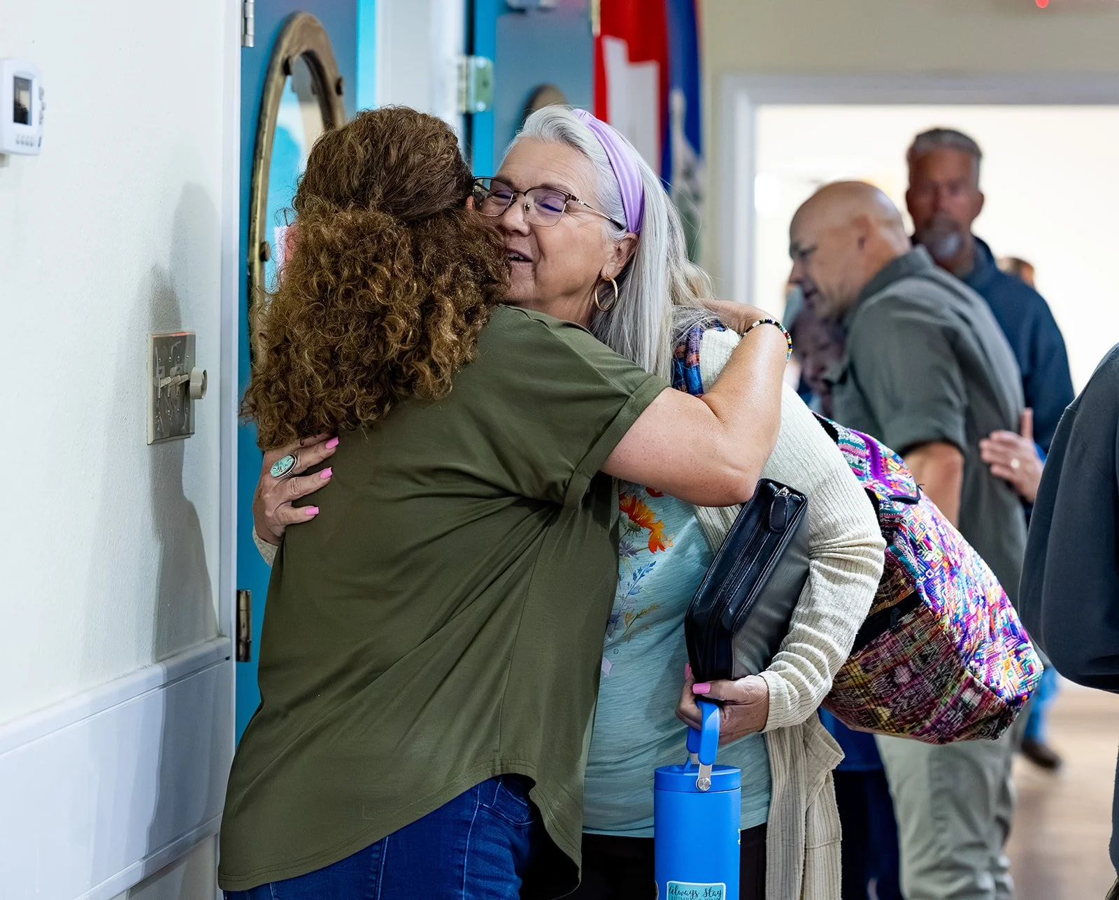 Two women hugging at what appears to be a social gathering or event, with other people in the background.