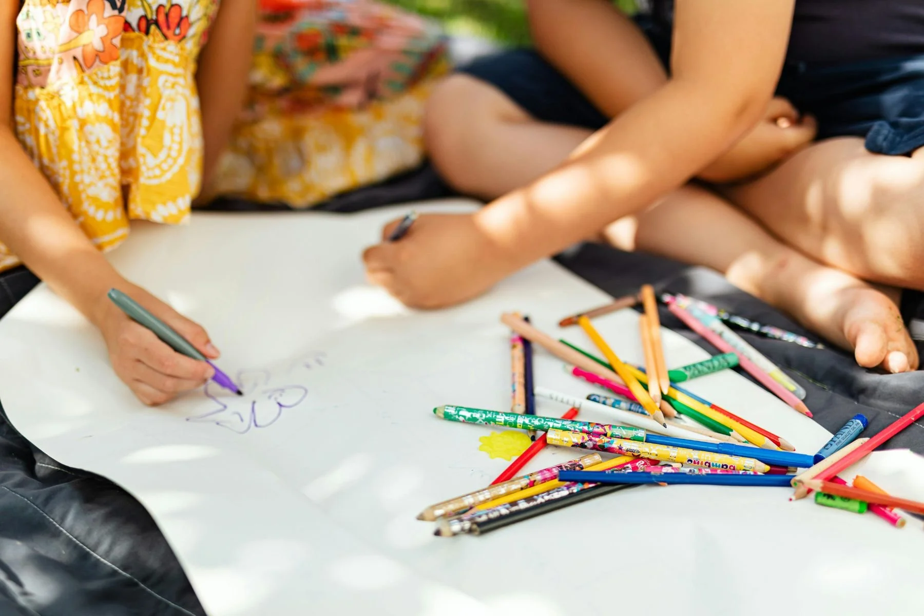 Children sitting on a white paper surface drawing with colored markers and pencils, with various art supplies scattered around.