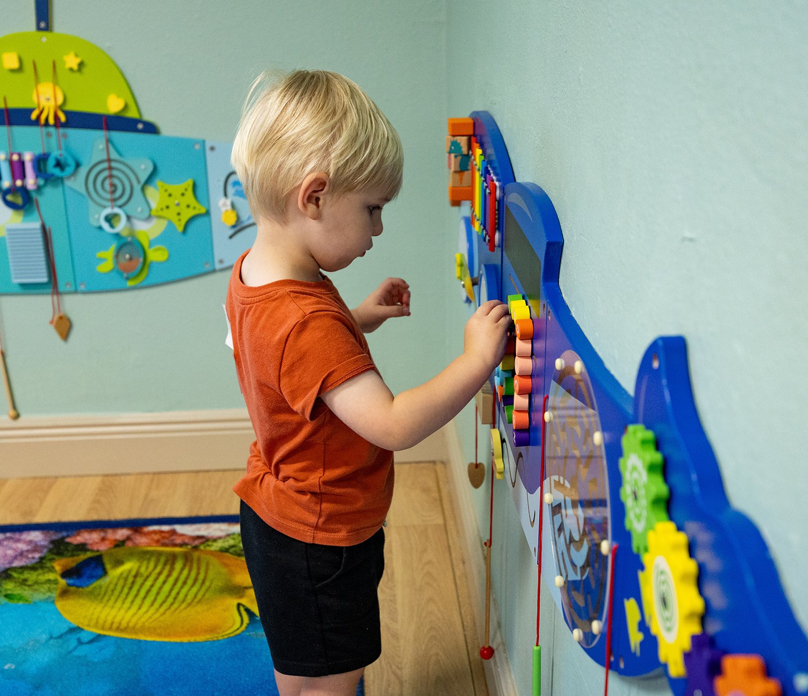 Young boy placing magnetic shapes on a blue wall-mounted artwork in a church nursery playroom with a colorful fish rug on hardwood floor and sea-themed decorations.