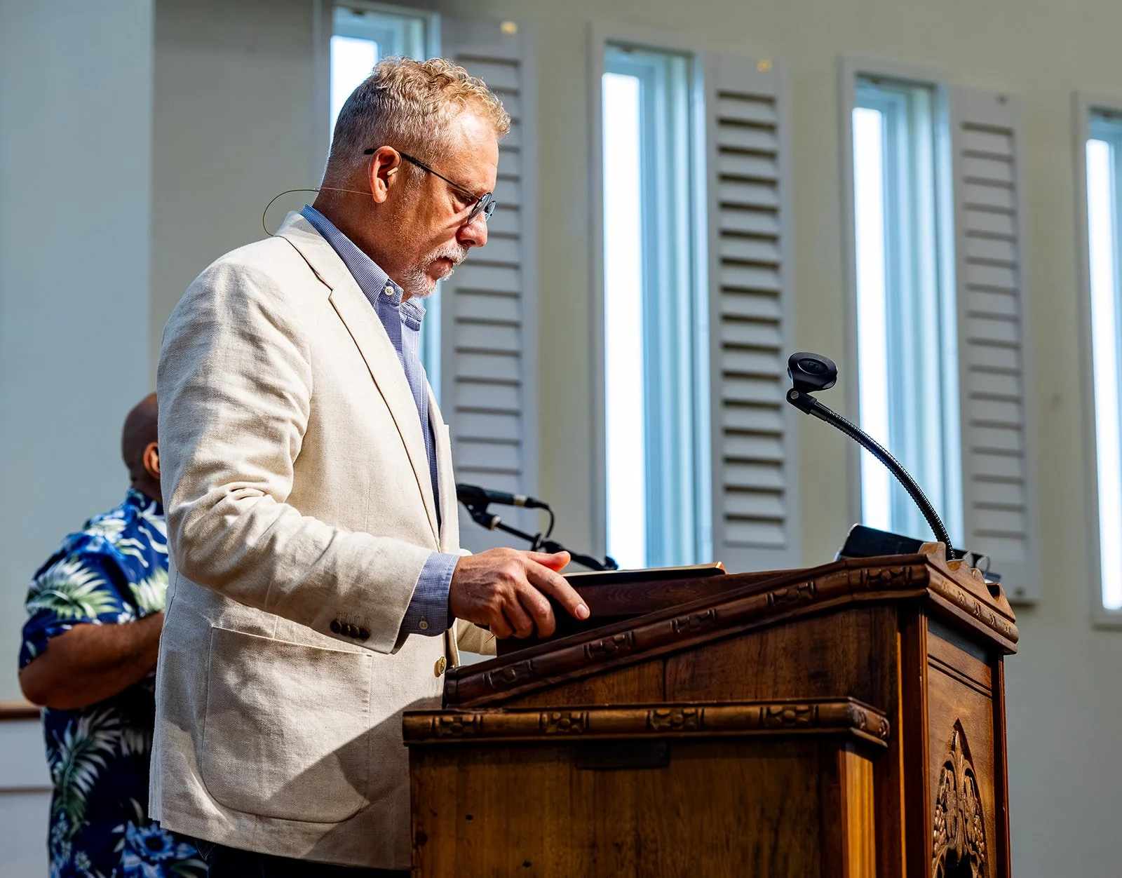 Pastor Thom Harrison of Padre Island Baptist Church, in a beige suit, standing at a wooden podium reading or speaking in a room with tall windows and white walls. Another person is partially visible behind him, wearing a Hawaiian shirt.