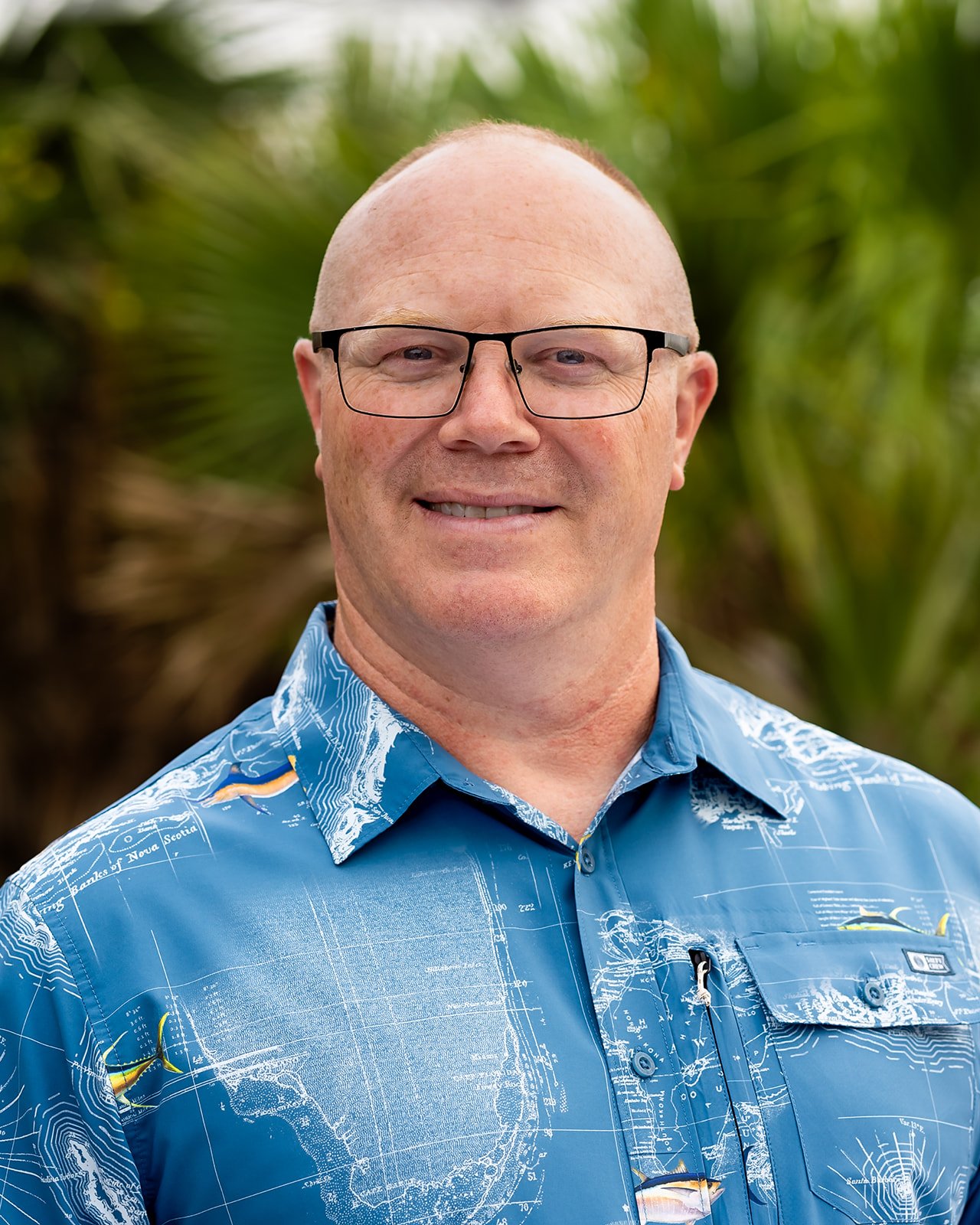 Pastor Steve of Padre Island Baptist Church wearing glasses and a blue Hawaiian shirt with a map design, standing outdoors with green plants in the background.