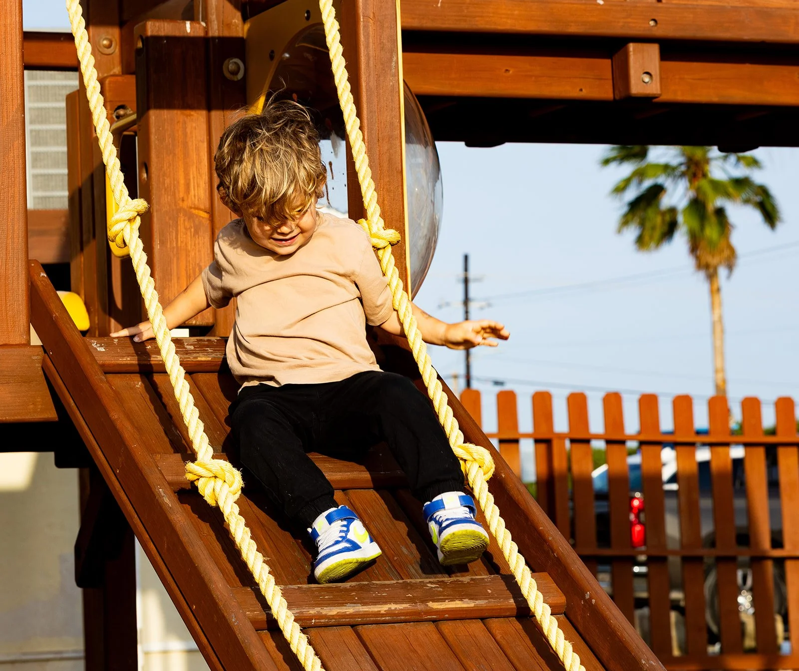 A young boy with messy hair wearing a beige shirt, black pants, and colorful sneakers, is sitting on a wooden slide in a playground, holding onto thick yellow ropes on either side.