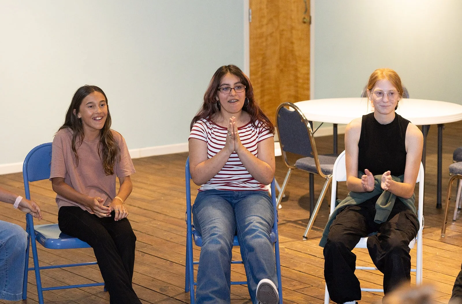 Three young women sitting in chairs inside a room with wooden floors and light green walls, smiling and clapping, during a group activity or gathering.