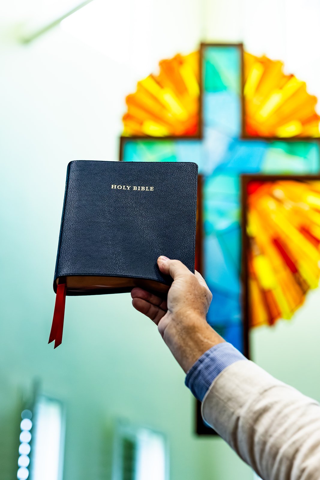 A person holding a black Holy Bible with a red ribbon bookmark against a background of colorful stained glass in the shape of a cross.