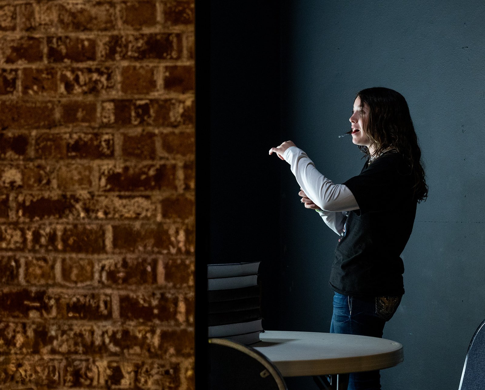 A woman standing beside a dark wall, smoking a cigarette with a shadowed brick wall on the left side of the image.
