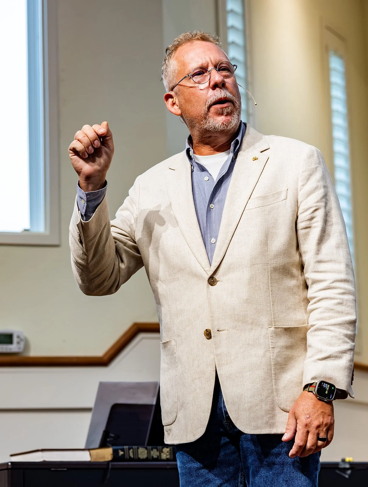 Pastor Thom Harrison of Padre Island Baptist Church in Corpus Christi, TX, is wearing a cream blazer, blue checkered shirt, and jeans. He appears to be giving a semon or in a church sanctuary with light-colored walls and windows.
