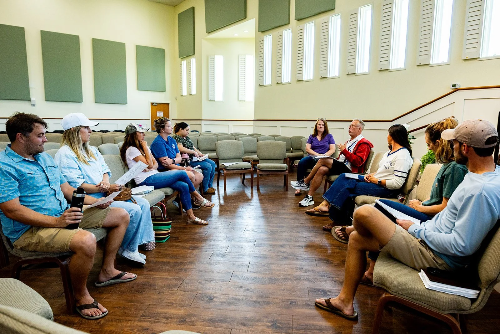 A group of diverse people sitting in a semi-circle in a large room with high ceilings, wood floors, and tall windows, participating in a discussion or meeting.