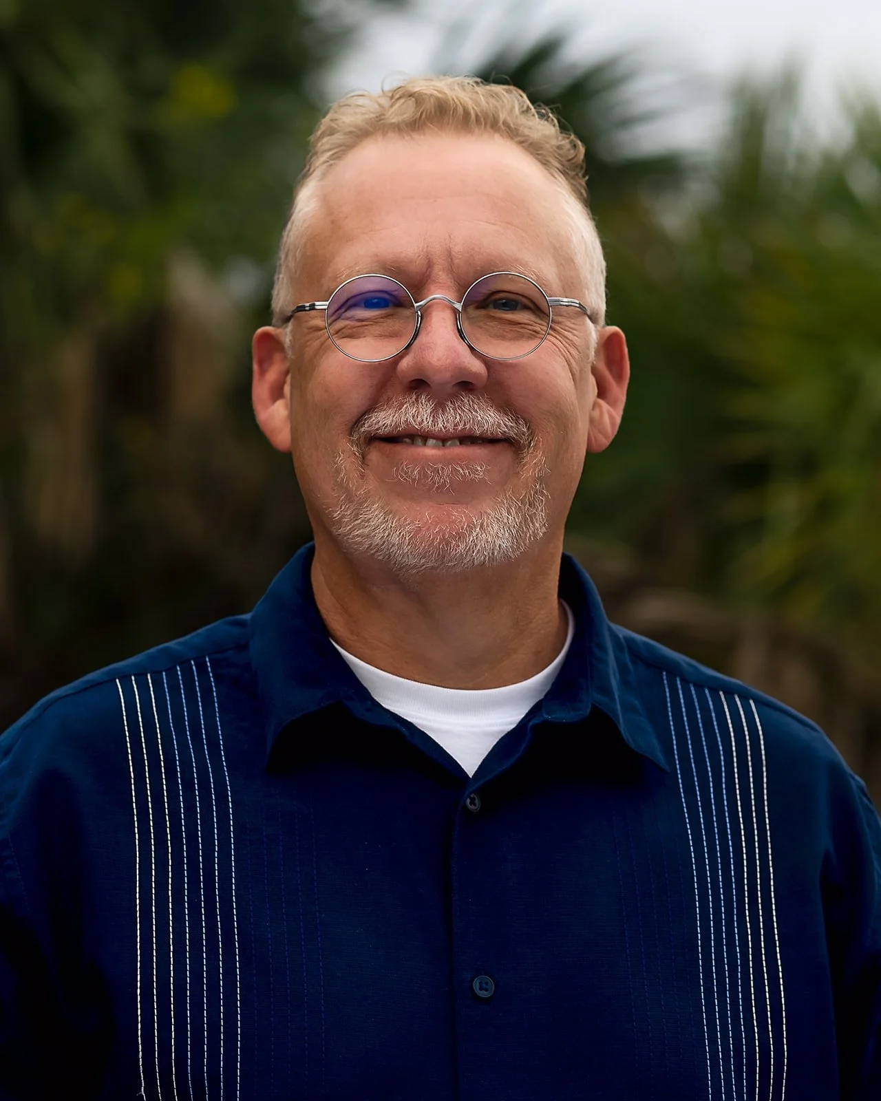 Pastor Thom Harrison of Padre Island Baptist Church in Corpus Christi, TX, with glasses and a goatee, wearing a blue shirt with white vertical stripes, standing outdoors with a blurred green background.