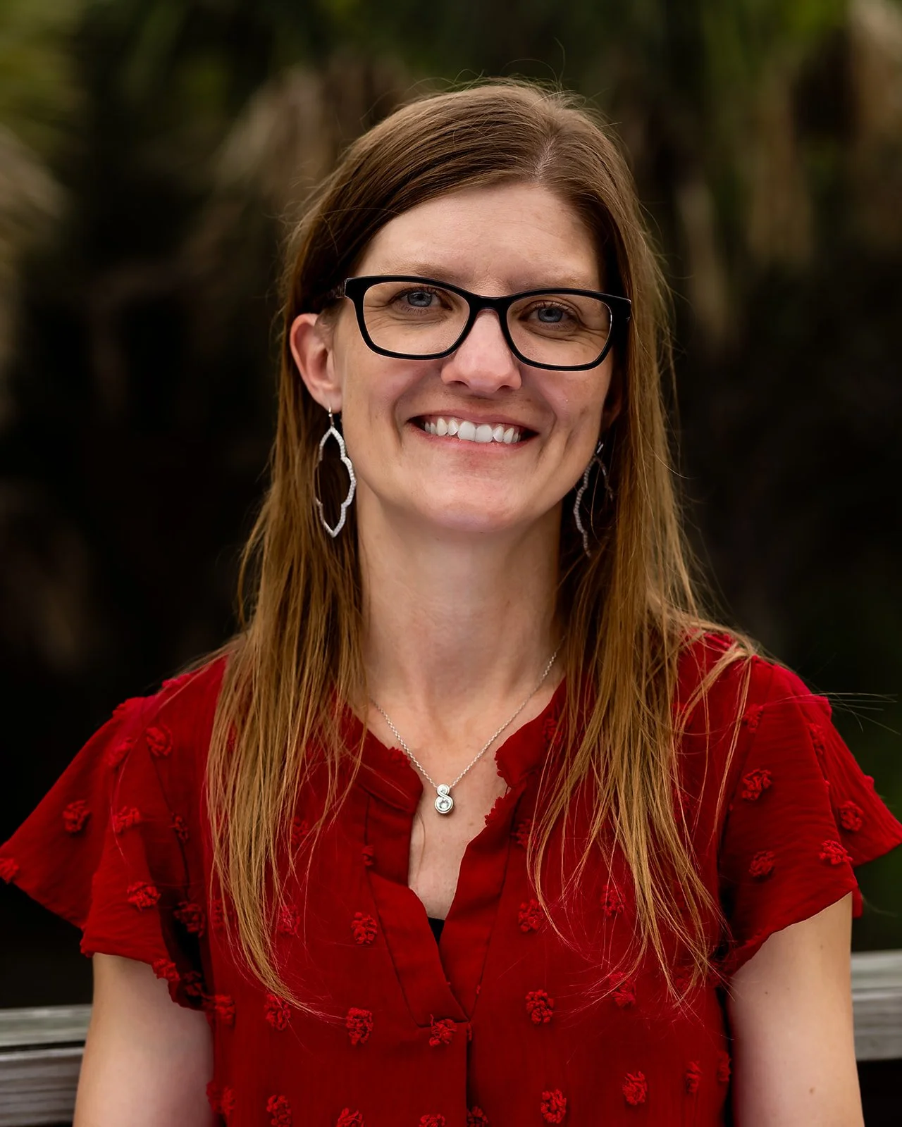 PBIC Staff member with long red hair wearing black glasses, earrings, a necklace with an S pendant, and a red blouse with textured embroidery, smiling at the camera.